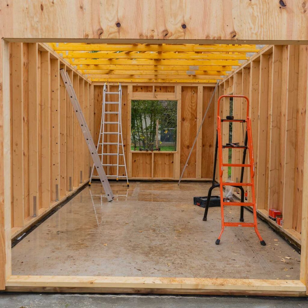Construction garage : ossature bois neuve et chantier intérieur. Intérieur de l'ossature bois d'un garage. Le sol en béton humide reflète les montants et deux échelles (argentée et orange) sur le chantier.