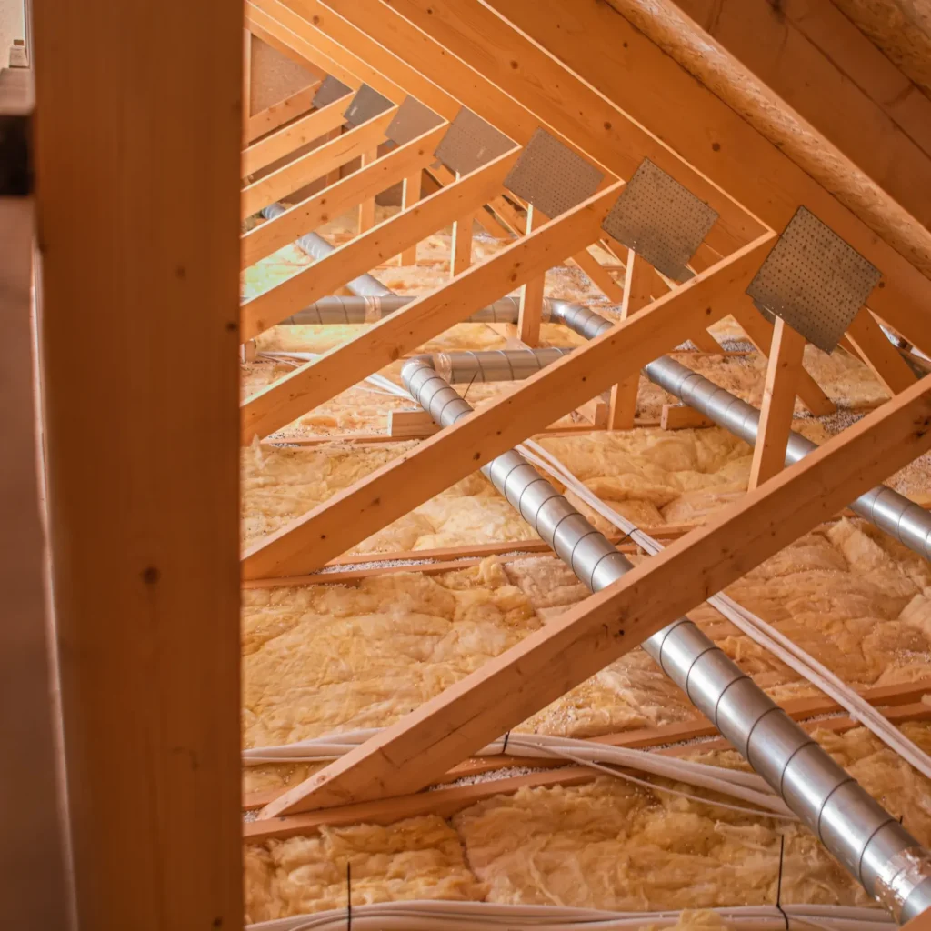 Isolation combles: charpente bois et conduits de ventilation Charpente de combles en bois (fermettes) avec isolation thermique en fibre de verre. Conduits de ventilation métalliques rigides et tuyaux PEX installés.
