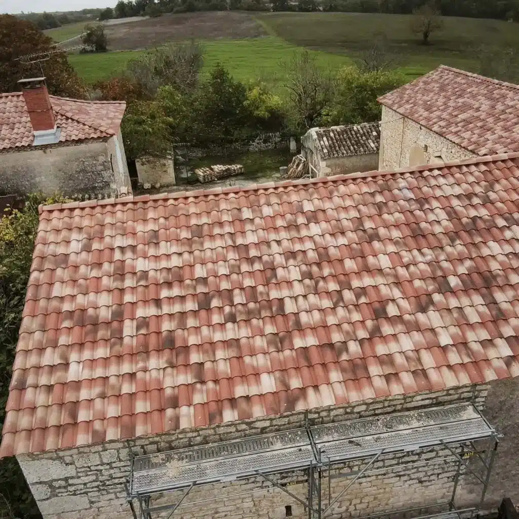 Rénovation de toiture : belles tuiles rouges sur maison ancienne. Toiture neuve en tuiles canal rouges et ocre sur maison en pierre. Vue aérienne de la restauration avec échafaudage et champs en arrière-plan.